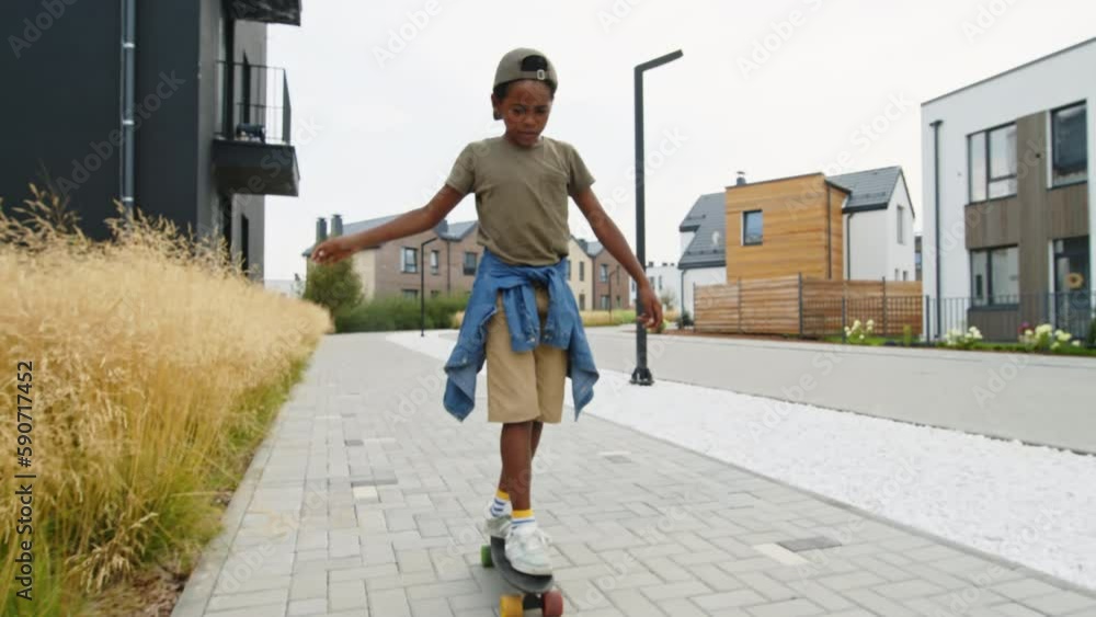 Full shot of happy African American child in casual clothes riding on skateboard on pavement near modern residential houses, trying to keep balance and smiling Full shot of happy African American child in casual clothes riding on skateboard on pavement near modern residential houses, trying to keep balance and smiling
