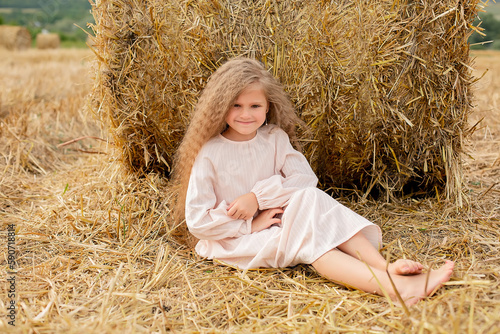 a beautiful blonde girl with long hair in a linen dress is sitting by a haystack