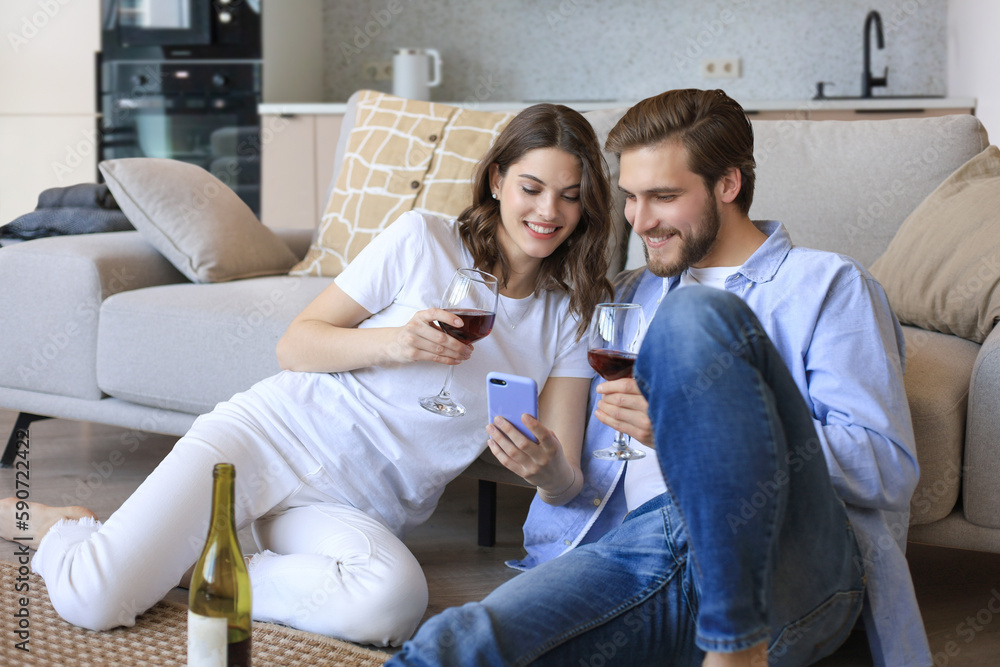 Happy couple sitting, relaxing on floor in living room, drinking red wine. Smiling young husband and wife rest at home enjoy romantic date on family weekend together Happy couple sitting, relaxing on floor in living room, drinking red wine. Smiling young husband and wife rest at home enjoy romantic date on family weekend together