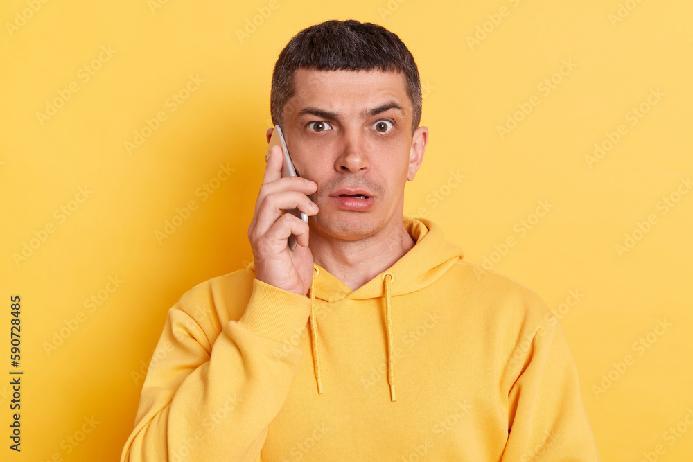 Fototapeta premium Shocked scared man wearing casual hoodie talking on smart phone hearing surprised news looking at camera with big eyes posing isolated over yellow background.