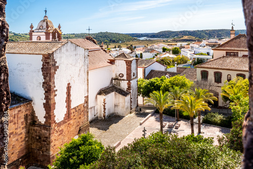 From castle of Silves, the Silves cathedral and its lateral square can be seen in front of Cidade de Silves rooftops, with palm trees and lush surroundings leading to Arade river under a sunny sky.