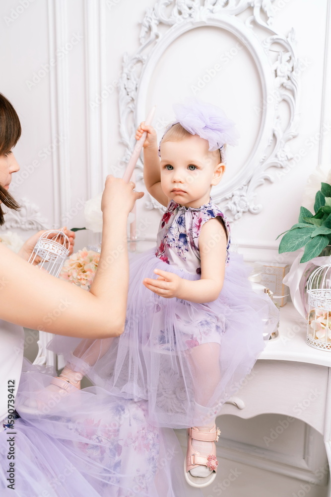 Fototapeta premium Baby girl elegant dress. A one-year-old girl in a puffy dress and a cute bow poses against the backdrop of a bright room with a dressing table and flowers.