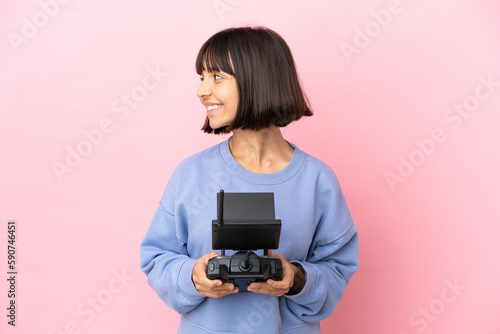 Young mixed race woman holding a drone remote control isolated on pink background looking side