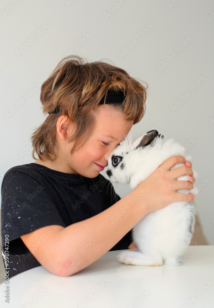 a boy of eight years old in a black T-shirt hugs a white rabbit, a child and a pet at home, preparing for Easter. The boys are playing in the living room