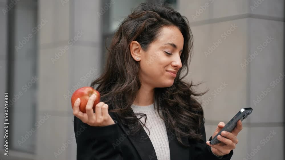 Pretty Caucasian lady looking at phone screen while eating an apple outdoors. Woman checking e-mail while working with smartphone. Technology, digital concept. Real time Pretty Caucasian lady looking at phone screen while eating an apple outdoors. Woman checking e-mail while working with smartphone. Technology, digital concept. Real time