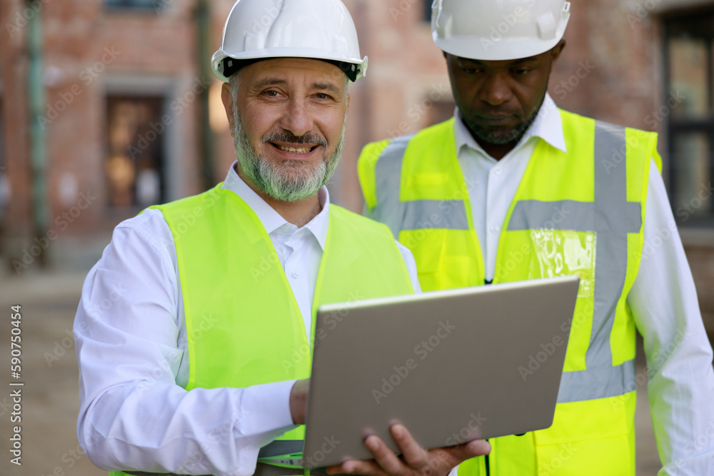 Portrait ofthe two builders together standing, looking at construction area with wireless laptop working on the project. Concept of business meeting and building process. Portrait ofthe two builders together standing, looking at construction area with wireless laptop working on the project. Concept of business meeting and building process.