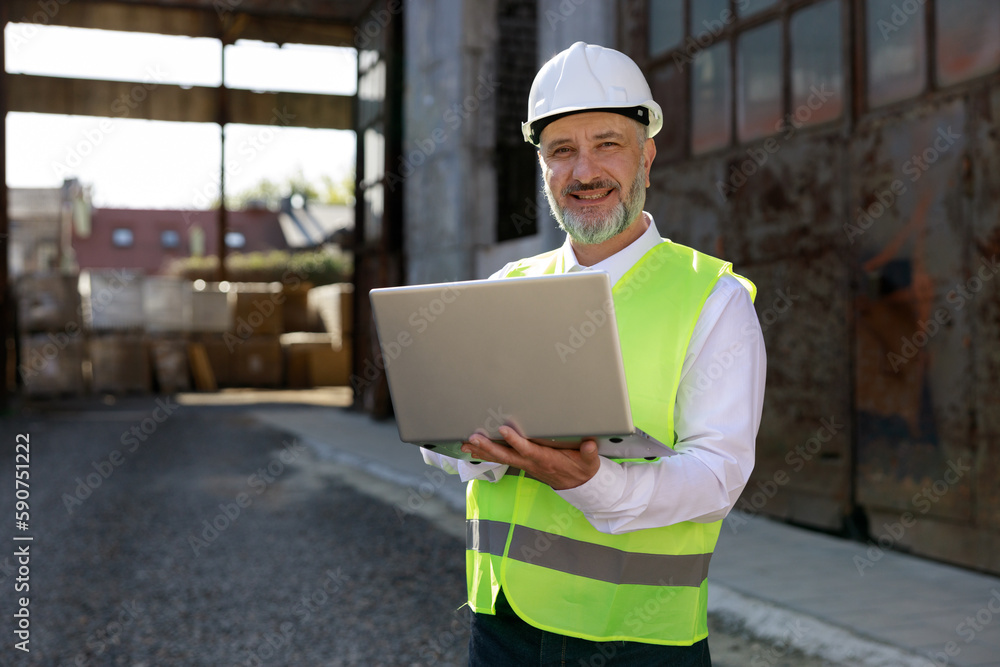 Portrait of the man builder in helmet and vest using wireless laptop for improving blueprints while standing at construction site. Concentrated bearded maleplanning system of building structures. Portrait of the man builder in helmet and vest using wireless laptop for improving blueprints while standing at construction site. Concentrated bearded maleplanning system of building structures.