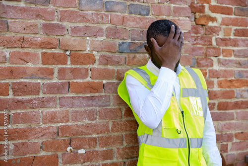 African american architect holding hands on head while suffering from severe headache outdoors. Tired builder in uniform standing near brick wall after hard working day.