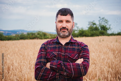 Arms crossed mature farmer man looking serious at camera standing at wheat field. Front view of folded agricultural rural worker with pensive attitude at harvest farm land. High quality photo