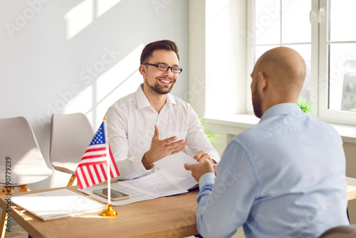 A young attractive man sitting in the office of the US public services or embassy with immigration application and having consular visa interview with US flag at the desk of official\'s workplace.