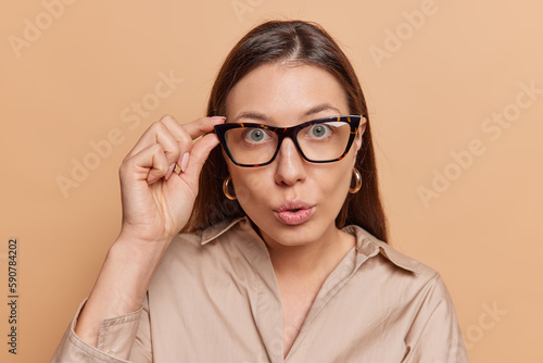 Portrait of surprised woman stares impressed at camera keeps hand on rim of spectacles wears shirt feels amazed reacts to shocking news isolated over brown background. Human reactions concept
