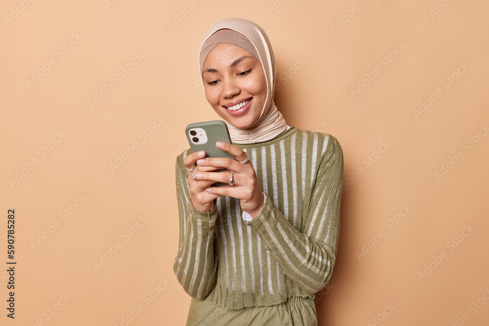 Waist up shot of pretty Islamic woman with cheerful expression wears hijab and jumper holds mobile phone sendstext messages smile gladfully isolated over brown background. Technology and religion Waist up shot of pretty Islamic woman with cheerful expression wears hijab and jumper holds mobile phone sendstext messages smile gladfully isolated over brown background. Technology and religion