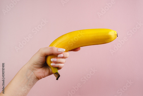 A young woman holds a banana in her hands on a pink background. Concept photo, female independence, feminism. Banana gun