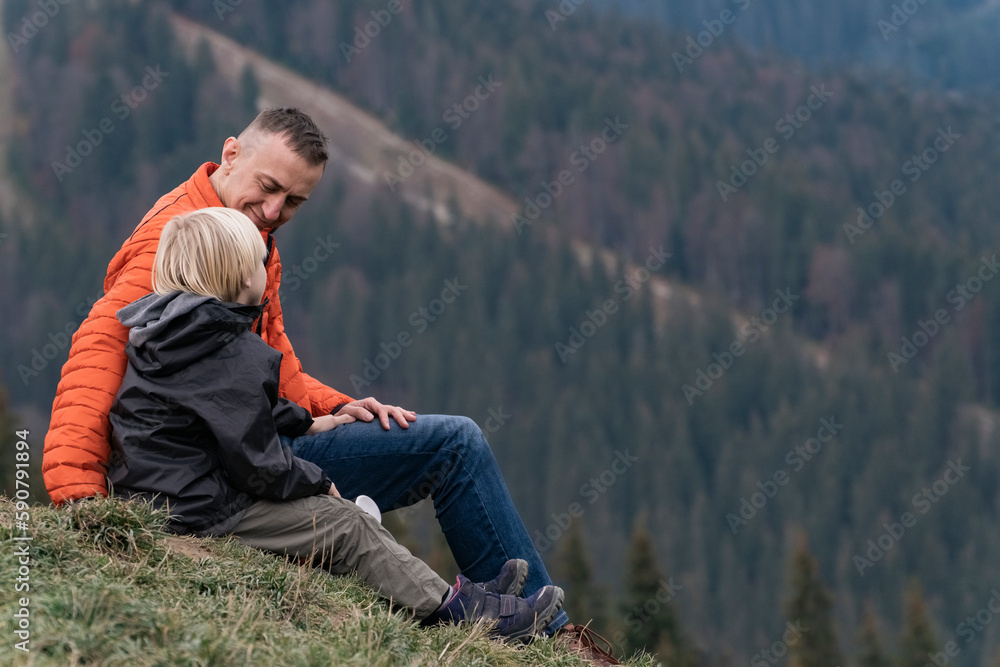 Father and son sit on hillside on the forest background. Weekend hike in mountains. Trekking and hiking with children. Father and son sit on hillside on the forest background. Weekend hike in mountains. Trekking and hiking with children.