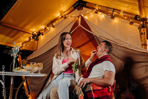 Happy lovely couple relaxing in glamping on summer evening and eating grapes near cozy bonfire. Luxury camping tent for outdoor recreation and recreation. Lifestyle concept © bondvit