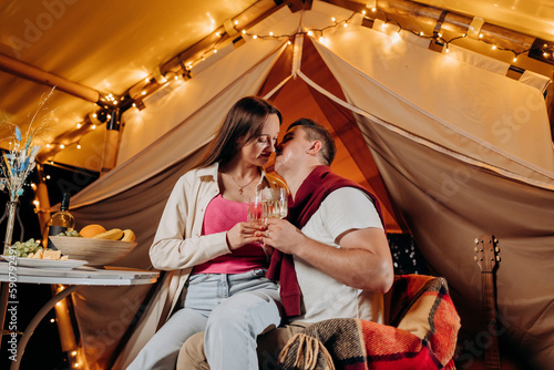 Happy lovely couple relaxing in glamping on summer evening and drinking wine near cozy bonfire. Luxury camping tent for outdoor recreation and recreation. Lifestyle concept © bondvit
