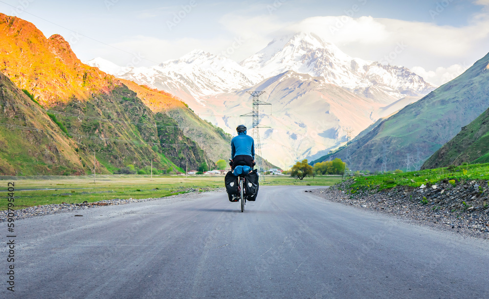 Male person in blue jacket cycling with touring bicycle surrounded by mountains and green summer nature. Tour around kazbegi national park. Georgia travel cycling holidays concept Male person in blue jacket cycling with touring bicycle surrounded by mountains and green summer nature. Tour around kazbegi national park. Georgia travel cycling holidays concept