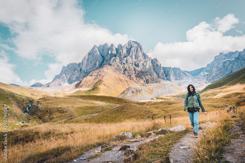 Front happy female caucasian woman hiker walks on pathway in Juta valley with Kazbegi caucasus mountains landscape. KAzbegi national park explore trekking concept