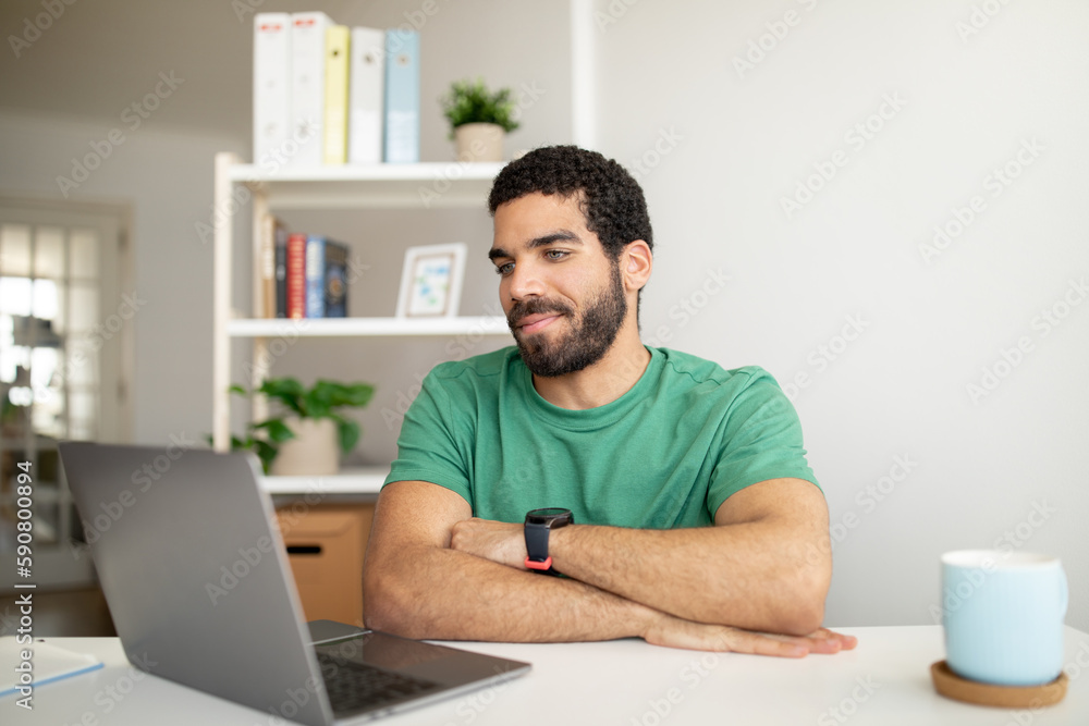 Smiling handsome millennial arabic man with beard has video call, working with laptop at workplace in room Smiling handsome millennial arabic man with beard has video call, working with laptop at workplace in room