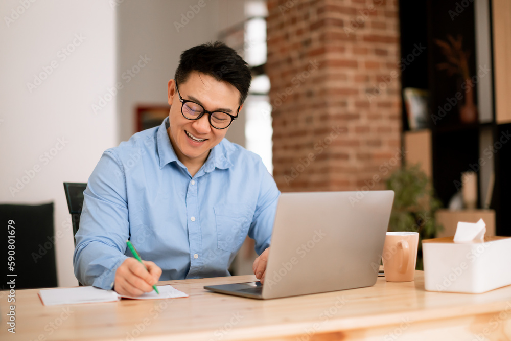 Happy asian man making notes, analyzing data for investment at workplace with laptop computer in home office interior Happy asian man making notes, analyzing data for investment at workplace with laptop computer in home office interior