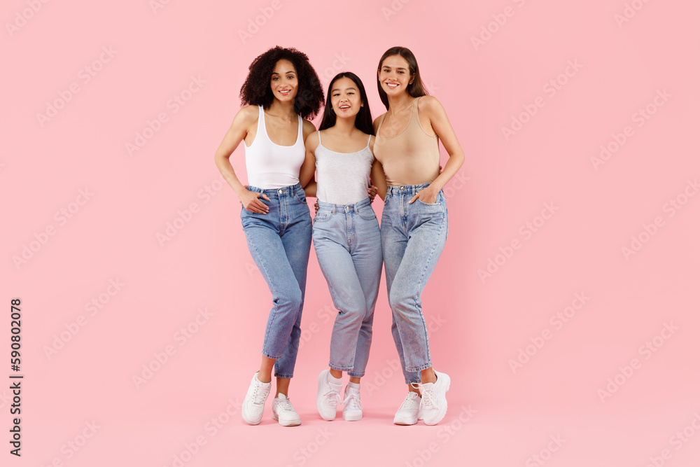Fototapeta premium Happy three multiracial girls embracing and smiling to camera, posing in studio over pink background, full body lenght