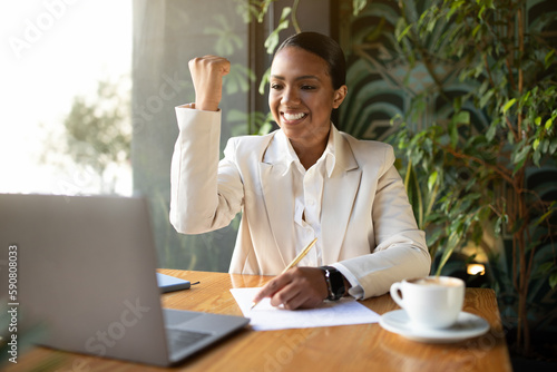 Happy millennial african american businesswoman in white suit working on laptop, making victory hand gesture