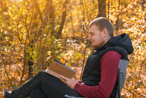 Young man is sitting on chair in the autumn forest and reading book. Mental recreation in nature. Quiet country life. Escapism. Digital detoxification. Concept of reading a book in the autumn park.
