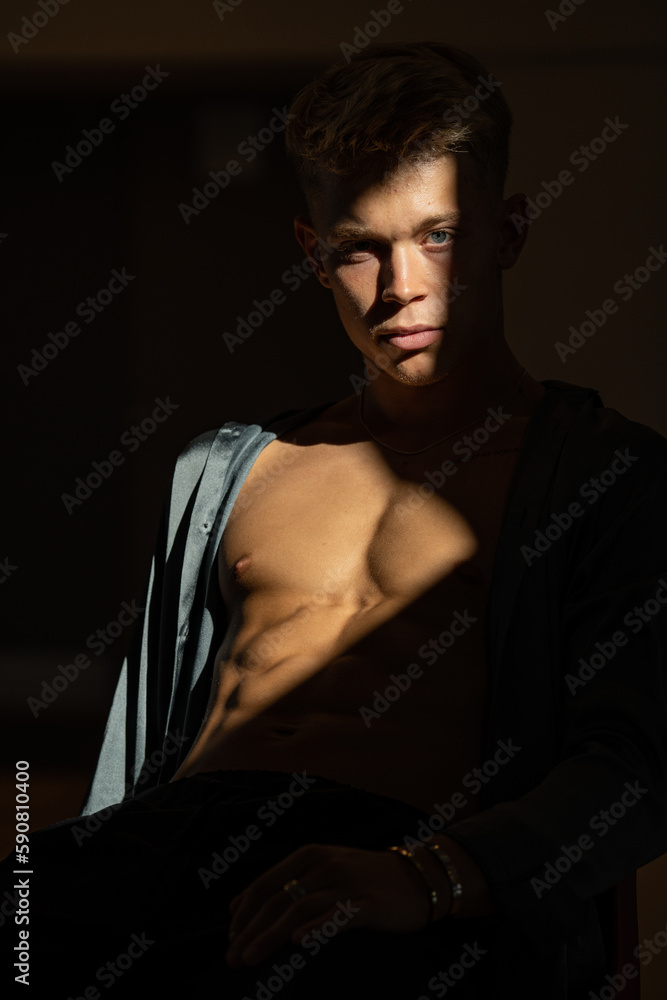 Fototapeta premium A muscular man in a gray satin shirt sitting on a dark background with falling sunlight