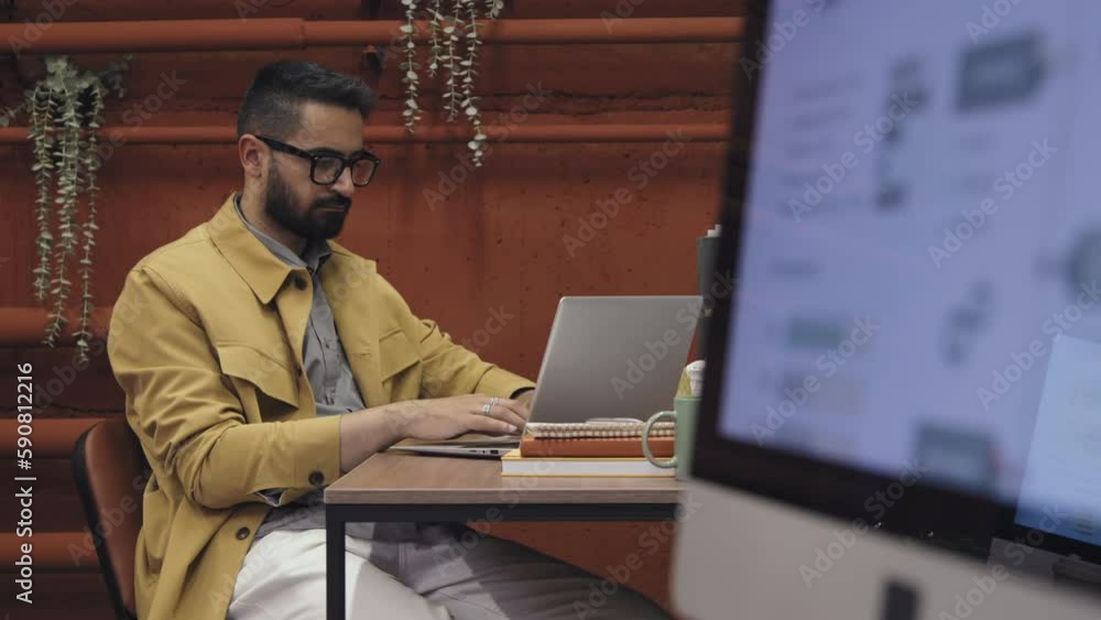 Selective focus shot of Middle Eastern male employee with beard, in glasses sitting at desk, typing on laptop in shared loft-style office, and presentation slide on screen in blurred foreground Selective focus shot of Middle Eastern male employee with beard, in glasses sitting at desk, typing on laptop in shared loft-style office, and presentation slide on screen in blurred foreground