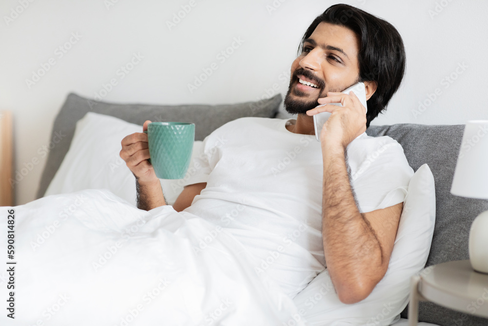 Smiling millennial arabic guy with beard lies on bed with cup of coffee calls by smartphone in bedroom Smiling millennial arabic guy with beard lies on bed with cup of coffee calls by smartphone in bedroom