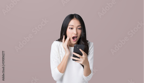 Pretty Asian woman looking at smartphone screen feeling surprise on isolated brown background
