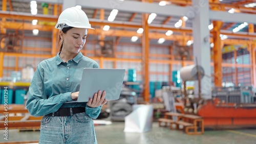 Professional engineer woman manager leader holding and typing on laptop wearing helmet standing at manufacturing factory recheck stock of production in workplace area