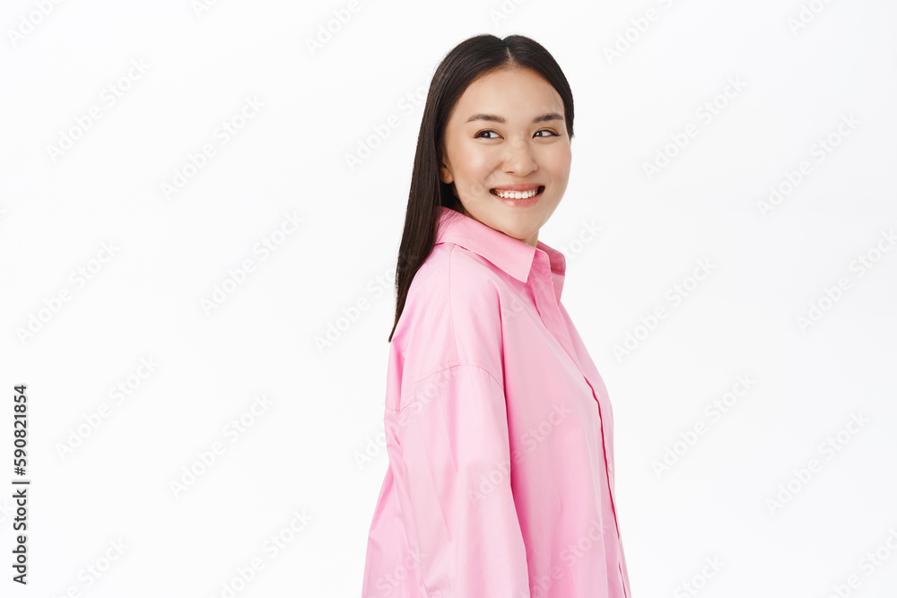 Smiling young asian girl looks behind her shoulder, stands confident and happy, wears pink shirt, white background Smiling young asian girl looks behind her shoulder, stands confident and happy, wears pink shirt, white background