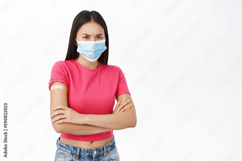 Upset asian girl in medical face mask, has band aid on shoulder from covid-19 vaccine, looking disappointed, standing over white background Upset asian girl in medical face mask, has band aid on shoulder from covid-19 vaccine, looking disappointed, standing over white background