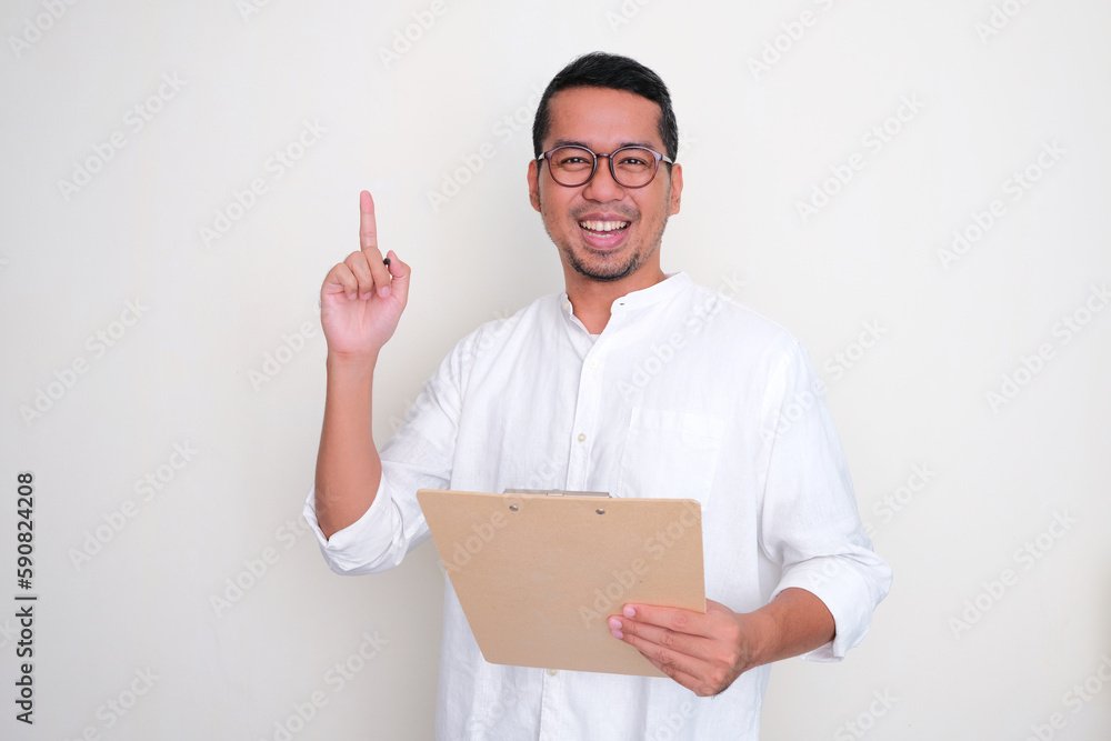 Adult Asian man smiling happy and pointing finger up while holding a clipboard Adult Asian man smiling happy and pointing finger up while holding a clipboard