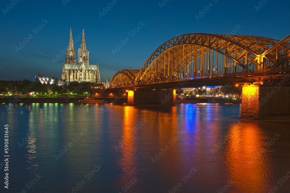 Fototapeta premium Eee foto in de avond met een lange sluitertijd van de oude Hohenzollernbrücke over de Rijn in Keulen. Op de achtergod is de mooie verlichte gotische kathedraal de Dom te zien bij een blauwe lucht.