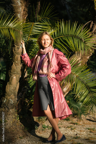 A woman in a fancy raincoat stands near a palm tree.
