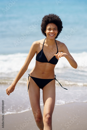 Joyful black woman in bikini walking along seaside