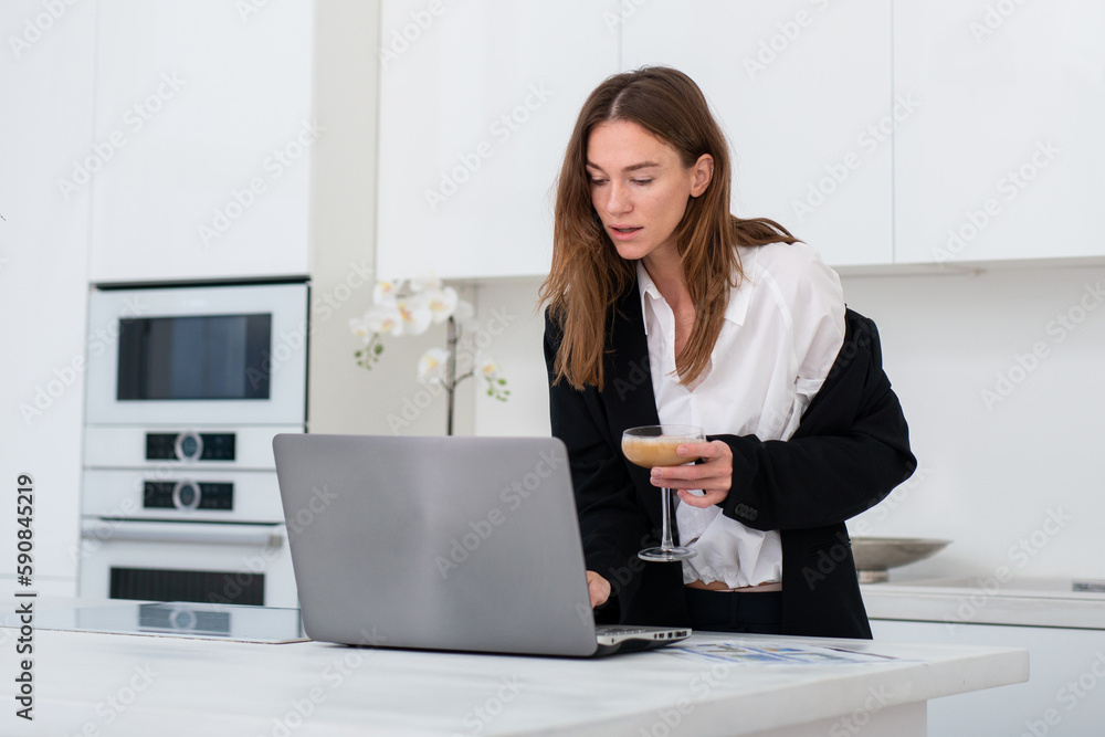 Relaxed woman at home in the kitchen after work drinking from a glass in front of her laptop computer