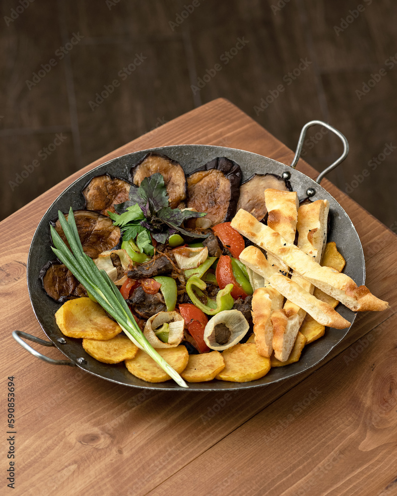 Fried vegetables with pita bread and green onions in metal brazier. Baked eggplant, tomatoes, peppers, potatoes. Wooden background. View from above. Copy space. 