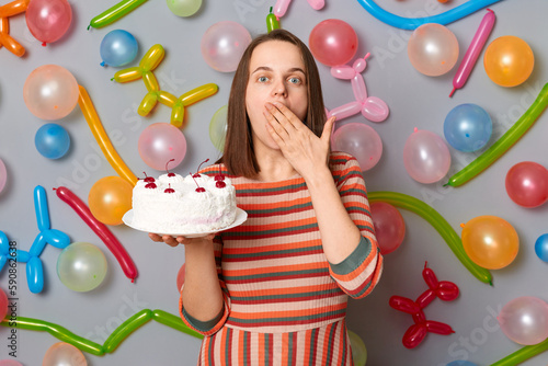 Amazed surprised woman with brown hair wearing striped dress holding cake covering mouth with hands sees shocked birthday present standing against gray wall decorated with colorful balloons.
