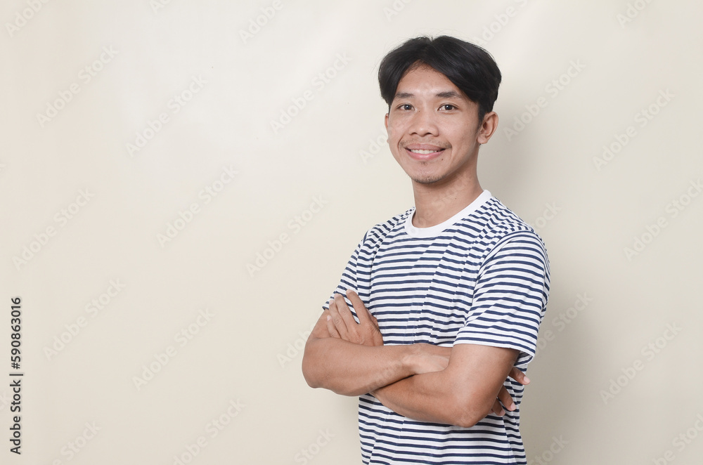 young asian man in striped t-shirt standing with arms crossed over isolated background and smiling young asian man in striped t-shirt standing with arms crossed over isolated background and smiling