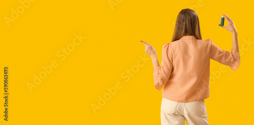 Young woman with inhaler pointing at something on yellow background with space for text, back view