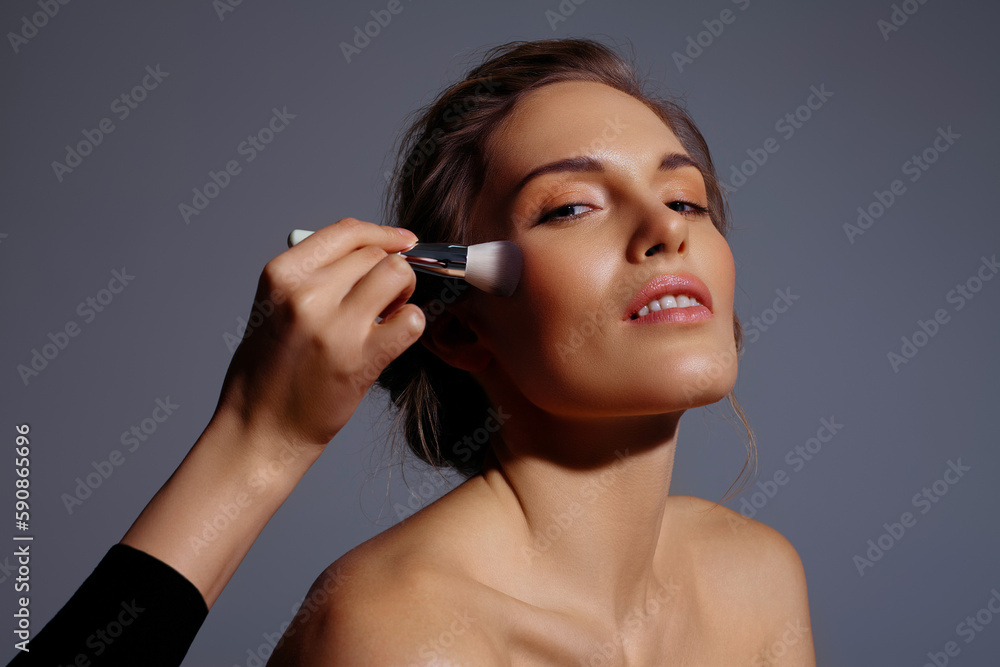 Correcting makeup with brush on face of beautiful blonde woman on gray background Correcting makeup with brush on face of beautiful blonde woman on gray background