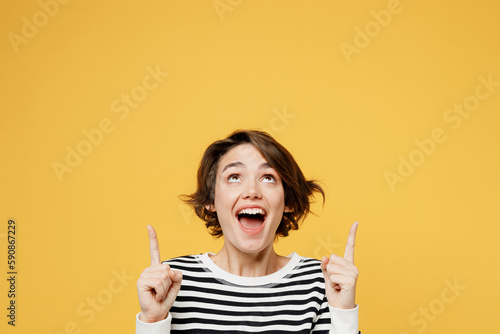 Young excited fun woman wear casual striped black and white shirt point index finger overhead indicate on workspace area copy space mock up isolated on plain yellow color background studio portrait.