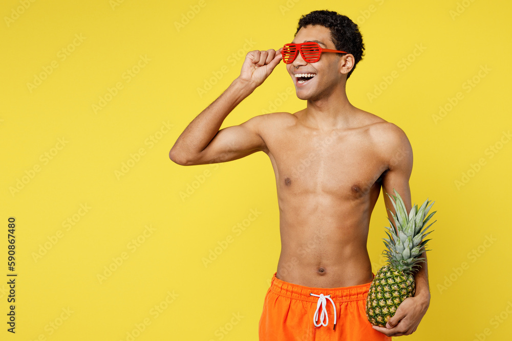 Handsome happy smiling young man wear orange shorts swimsuit hold in hand pineapple looing aside relax near hotel pool isolated on plain yellow background. Summer vacation sea rest sun tan concept. Handsome happy smiling young man wear orange shorts swimsuit hold in hand pineapple looing aside relax near hotel pool isolated on plain yellow background. Summer vacation sea rest sun tan concept.