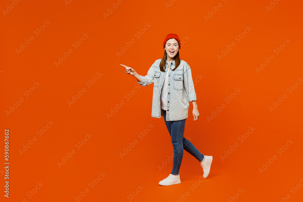 Sideways young smiling surprised shocked woman wearing denim shirt white t-shirt red hat look camera walking going point index finger aside on area isolated on plain orange background studio portrait. Sideways young smiling surprised shocked woman wearing denim shirt white t-shirt red hat look camera walking going point index finger aside on area isolated on plain orange background studio portrait.