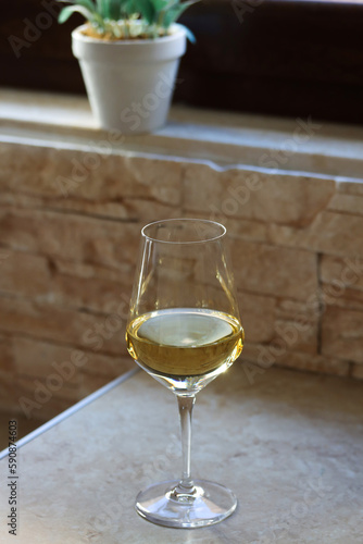 A glass of white wine on the kitchen table in front of the window with a flower on the back on a sunny day. Close up of a shining glass of white wine on a table with copy space for text