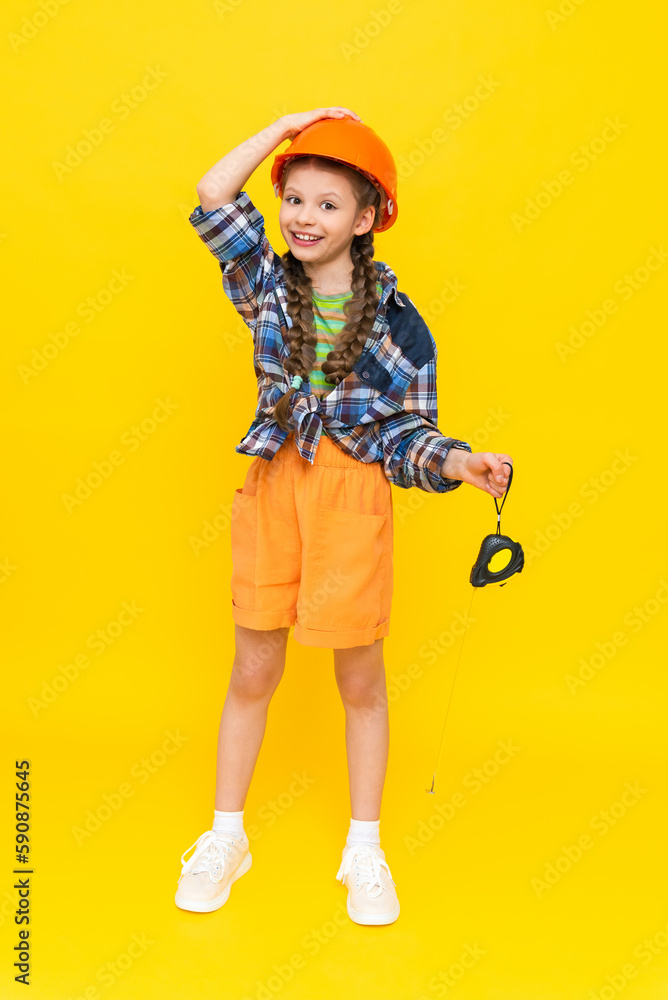 A little girl in a helmet holds a construction tape measure. A child builder in a full-length plaid shirt. The concept of renovation in the children's room. Yellow isolated background. A little girl in a helmet holds a construction tape measure. A child builder in a full-length plaid shirt. The concept of renovation in the children's room. Yellow isolated background.