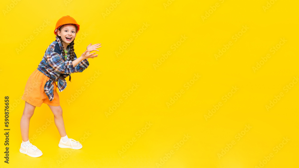 Naklejka premium A child in a construction helmet points to your advertisement. Happy little teen girl in full height. A place to advertise. Yellow isolated background. Copy space. Banner.